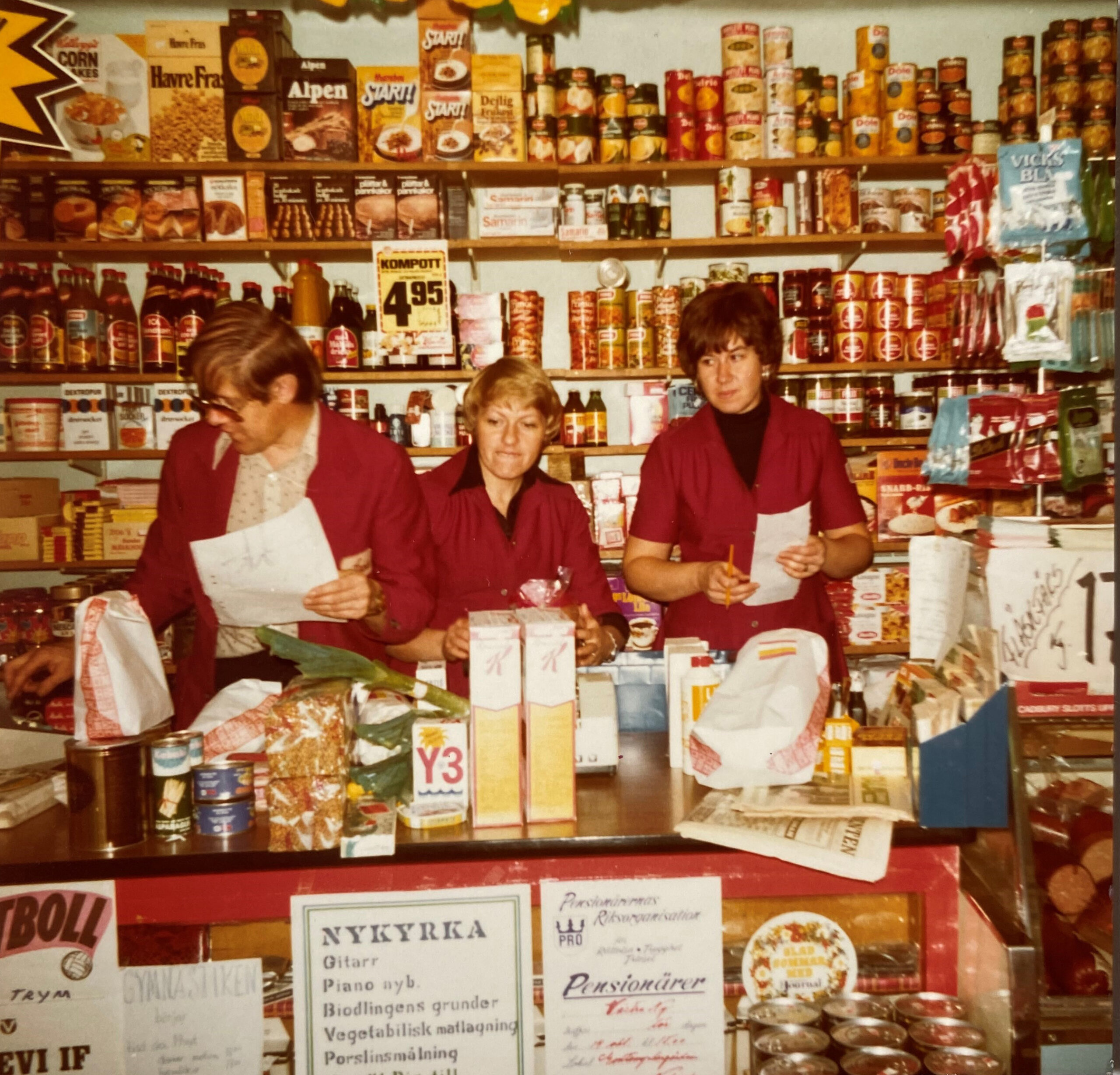 Ett foto från 70-talet på tre personer som står bakom en affärsdisk. Hyllorna på väggen är fulla av varor.
