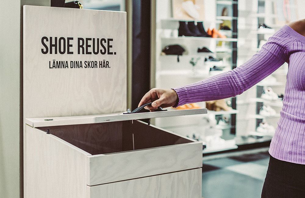 Kvinna i lila tröja lägger ett par skor i en låda med texten "Shoe Reuse".