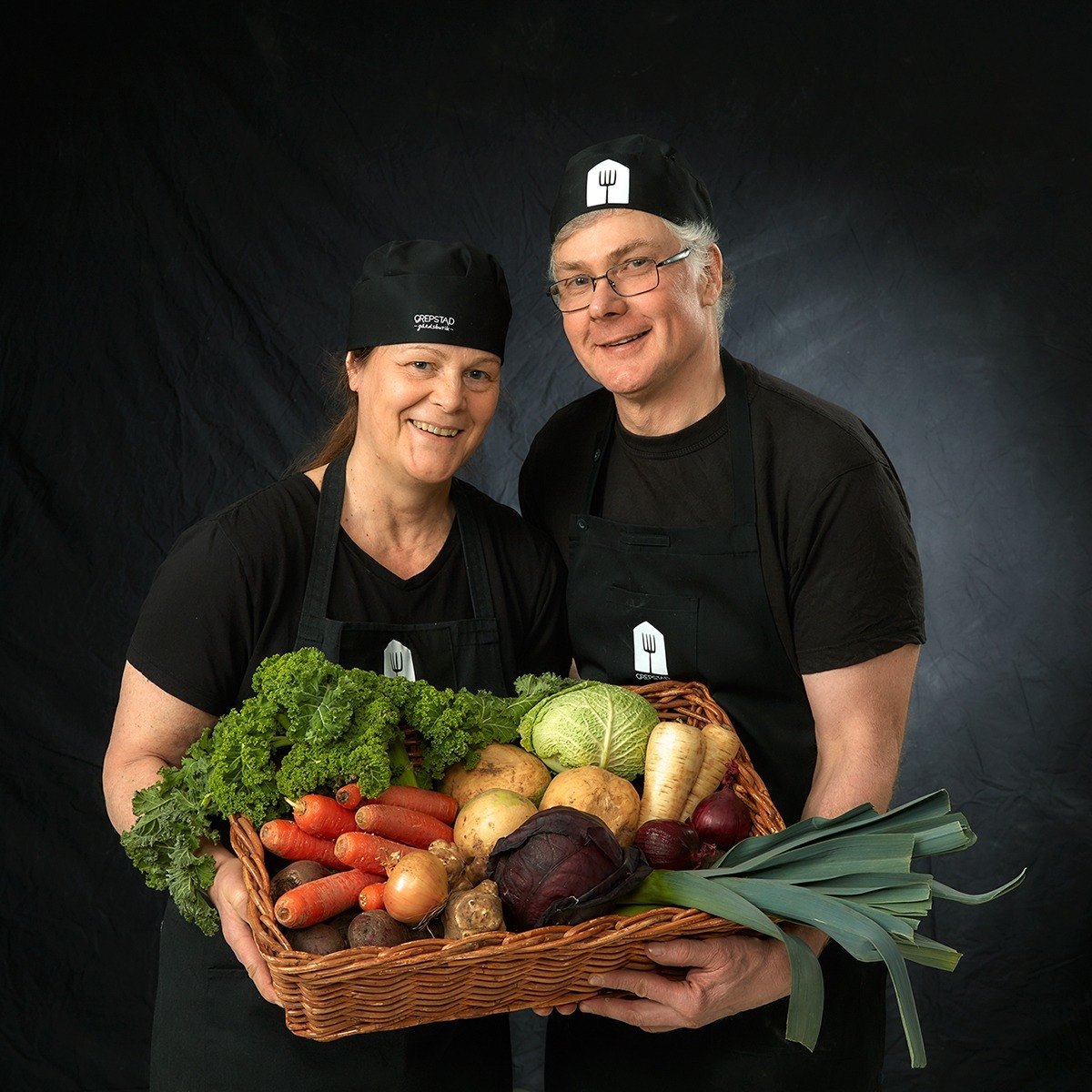Porträtt av man och kvinna i svarta kläder som har famnarna fulla av grönsaker.