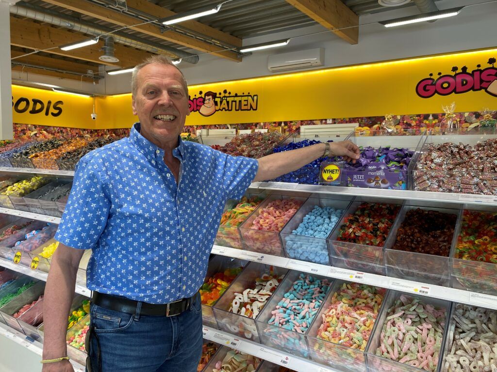 En leende man står i en godisbutik, klädd i jeans och blå skjorta tar en lilafärgad pralin.