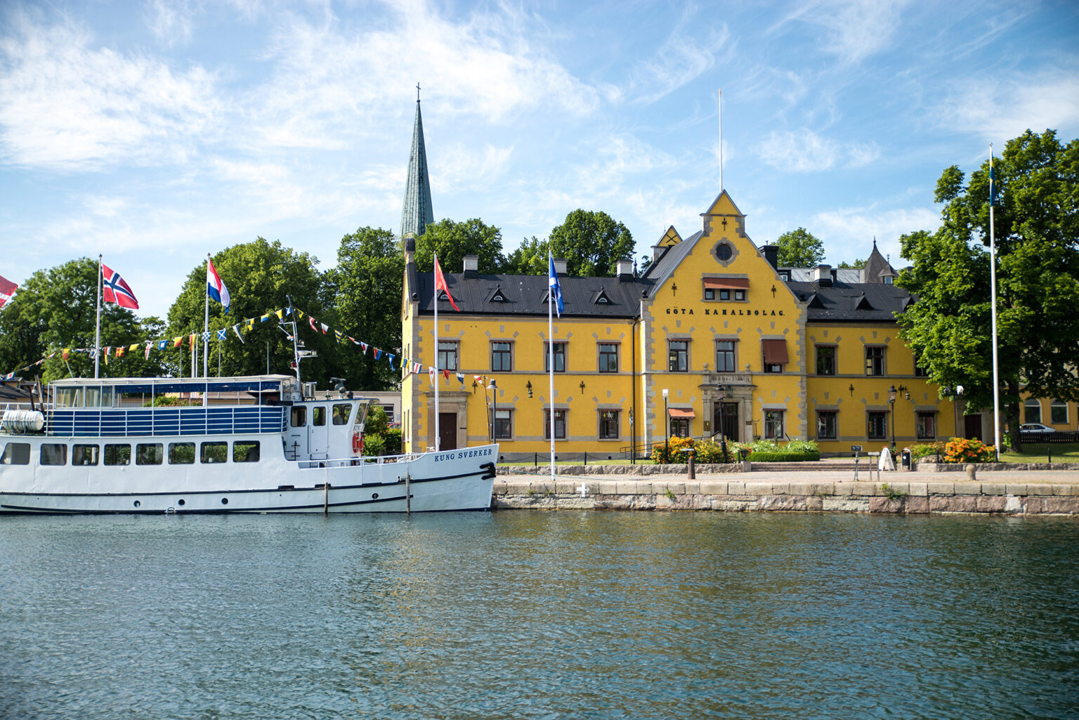 Det stora gula kanalbolagshuset i Motala hamn syns intill en blå himmel och båten Kung Sverker.